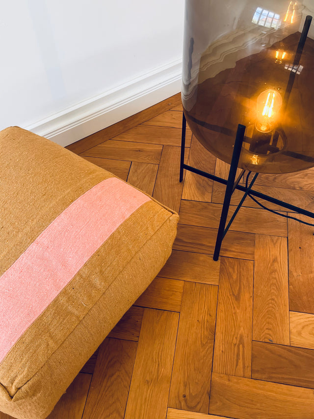 Farbenfrohes Bodenkissen von LEY auf Fischgrätparkett neben Designerlampe.