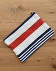 Die LEY! Tasche - Stripe Line "Parosa" (20x30cm) aus der Stripe Edition zeichnet sich durch kräftige und dünne Querstreifen in Rot, Weiß und Marineblau aus, die auf einer hellen Holzoberfläche abgebildet sind.