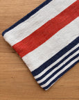 Ein gefaltetes Tuch aus der Tasche - Stripe Line "Parosa" (20x30cm) von LEY! mit kräftigen roten, weißen und blauen Streifen ruht auf Holz - ein idealer stilvoller Akzent für Ihre Baumwolltasche.