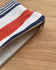 Die LEY! Tasche - Stripe Line "Parosa" (20x30cm), mit roten, weißen und blauen Streifen, liegt teilweise entpackt auf einer Holzunterlage.