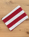 Die Tasche - Stripe Line "Symia" (20x30cm) von LEY! aus Baumwolle mit roten und weißen Streifen und Reißverschluss liegt auf hellem Holz - handgefertigte Qualität aus Marrakesch.