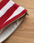 Die Tasche - Stripe Line "Symia" (20x30cm) von LEY! mit rot-weißen Streifen und beigem Reißverschluss liegt halb geöffnet auf heller Holzoberfläche.