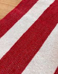 Die Nahaufnahme der LEY! Tasche - Stripe Line "Symia" (20x30cm) zeigt den gewebten Stoff mit abwechselnd roten und weißen Streifen, die die Textur und das Webmuster auf einer Holzoberfläche hervorheben.