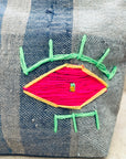 Eine Nahaufnahme des blau und grau gestreiften Stoffes der LEY! Tasche - Kosmetiktasche, mit einem bunten gestickten Auge, das gelb umrandet, mit leuchtend rosa Faden gefüllt und mit grün gestickten Wimpern akzentuiert ist.