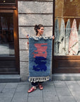 Eine Frau auf dem Bürgersteig hält vor einem Schaufenster einen „THE DANISH GIRL INTERIOR Berber-Teppich – Amani“ in den Händen.