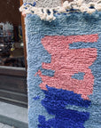 Ein blau-rosa Berber-Teppich – „Amani“ von THE DANISH GIRL INTERIOR – hängt draußen an einer grauen Wand neben einer Holztür.