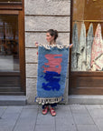 Eine Frau präsentiert den Berber-Teppich „THE DANISH GIRL INTERIOR“ – Amani, 0,60 x 1,10 m, mit blauen und rosa Mustern auf dem Bürgersteig.