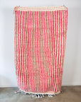 Der LEY! Berber Teppich - Pinq (1,20 x 2,00), handgewebt aus Schurwolle mit rosa-weißen Streifen und Fransen, hängt an einer weißen Wand - seine flauschige Struktur verleiht Charme und Wärme.