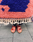 Eine Person in roten Sandalen steht neben dem Berber-Teppich „Farah“ (0,60 x 1,10 m) mit bunten Fransen von THE DANISH GIRL INTERIOR.