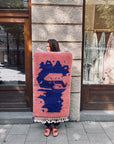 Eine Frau auf dem Bürgersteig hält den Berber-Teppich „THE DANISH GIRL INTERIOR“ – Farah, 0,60 x 1,10 m, in Rosa und Blau.