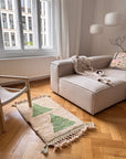 Ein gemütliches Wohnzimmer mit einem beigen Sofa, einem Holzstuhl, dem Berber-Teppich „Noor“ von THE DANISH GIRL INTERIOR und großen Fenstern.