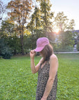 Eine Frau in einem Kleid mit Leopardenmuster und einer LEY! „Habibi“-Kappe in Blau steht auf einer Wiese, hinter den Bäumen scheint die Sonne.