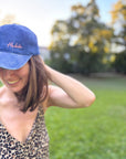 Eine lächelnde Frau in LEY! Mit einer „Habibi“-Kappe in Blau und einem Kleid mit Leopardenmuster steht sie auf einer Wiese.
