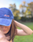Eine Frau trägt eine LEY! „Habibi“-Kappe in Blau mit verstellbarem Verschluss und steht im Freien zwischen verschwommenen Bäumen und Gras.