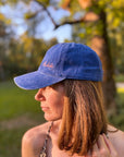 Eine Frau trägt die LEY! „Habibi“-Kappe in Blau im Freien, Sonnenlicht auf ihrem Gesicht und verschwommene Bäume im Hintergrund.