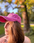 Eine Person mit langen braunen Haaren, die eine LEY! „Yalla Yalla“-Kappe in Lila trägt und im Freien vor einem verschwommenen grün-gelben Hintergrund steht.
