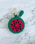 Der LEY! Schlüsselanhänger - Melone, ein kleines rundes Mini-Etui aus Kunstleder mit Reißverschluss in Wassermelonen-Design und goldener Befestigung, liegt auf einer hellen Marmoroberfläche.