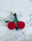 Der Schlüsselanhänger - Kirsche von LEY! zeigt zwei rote, runde Charms aus Mini Etui Kunstleder an grünen Stielen mit goldener Schlaufe, stilvoll auf Marmor präsentiert.