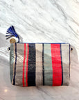 Die TASCHE von LEY! ist eine handgenähte Clutch mit blauen, roten und beigen Streifen, blauem Reißverschluss und Quaste mit Gold-Charme-ein Einzelstück, präsentiert auf weißem Marmor.