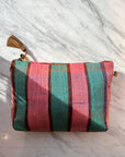 Die LEY! TASCHE ist eine kleine Stofftasche mit vertikalen rosa und blaugrünen Streifen, dünnen braunen Linien und einer braunen Quaste am Reißverschluss. Dieses einzigartige Stück wird auf einer weißen Marmoroberfläche im Sonnenlicht gezeigt.