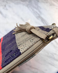 Nahaufnahme einer handgenähten LEY! TASCHE Clutch mit beigefarbenem Reißverschluss und Quaste, die mit einem Messingring versehen ist. Dieses Einzelstück zeigt beige, lila und rote Streifen auf einem marmorierten weißen Hintergrund.