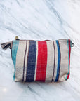 Die LEY! TASCHE ist eine rechteckige, gewebte Clutch mit blauen, roten, türkisfarbenen und braunen Längsstreifen und einem Reißverschluss mit Quasten, der auf Marmor ausgestellt ist. Dieses Einzelstück bringt einzigartiges Flair in Ihre handgenähte Taschensammlung.