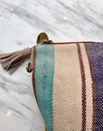 Eine Nahaufnahme der LEY! TASCHE Clutch mit Reißverschluss und beiger Quaste auf weißem Marmor, mit gewebten türkisfarbenen, beigen, burgunderroten und lila Streifen für einen einzigartigen Look.