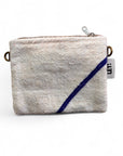 Eine kleine, rechteckige, strukturierte weiße Tasche (15x20cm) mit Reißverschluss, diagonalem blauen Streifen, Metallringen auf jeder Seite und einem weißen "LEY!"-Anhänger - eine Classic Line Tasche von LEY! aus Marrakesch.