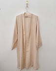 Der KIMONO - nude von BIRLIK in Einheitsgröße aus weicher, beiger Baumwolle mit langen Ärmeln und knielangem Schnitt hängt an einem pinken Drahtbügel vor einer weißen Wand.