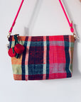 Eine rechteckige LEY! Handtasche mit einem lebhaften Karomuster in Rot, Pink, Grün, Blau und Gelb, mit einem pinkfarbenen Riemen und zwei Schlüsselanhängern - Kirsche, die am Reißverschluss hängen.