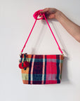 Eine Hand hält eine bunt karierte Handtasche mit einem leuchtend rosa Riemen und einer LEY! Schlüsselanhänger - Kirsche vor einem schlichten weißen Hintergrund.