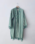 Der KIMONO - MEDITERANE - GRÜN - WEIß von THE DANISH GIRL INTERIOR mit Streifen, langen Ärmeln und Fransen hängt an einem Holzkleiderbügel vor einer schlichten weißen Wand.