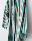 Der KIMONO - MEDITERANE - GRÜN - WEIß von THE DANISH GIRL INTERIOR mit langen Ärmeln und Fransen an den Bündchen besticht durch grüne und weiße Streifen für einen frischen, stilvollen Look vor weißer Wand.