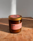 Eine braune Glaskerze mit goldenem Deckel steht auf Holz. Auf dem Etikett steht "LEY! Moments Duftkerze - Marrakesh Souks - Jasmin". Diese LEY! Duftkerze auf Sojabasis leuchtet, wenn das Sonnenlicht sanfte Schatten auf die Oberfläche wirft.