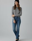 Eine Frau in einem grauen Langarmshirt, blauen Jeans und einer LEY!-Wollmütze „Saha“ in Schwarz von LEY! steht vor einem einfarbigen Hintergrund.
