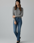 Eine Frau in einem grauen Langarmshirt, blauen Jeans und einer LEY!-Wollmütze „Saha“ in Schwarz von LEY! steht vor einem einfarbigen Hintergrund.