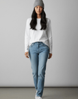 Eine Frau in einer beigen Unisex-Wollmütze „Saha“ von LEY!, einem weißen Langarmshirt und einer blauen Jeans steht vor einem einfarbigen Hintergrund.