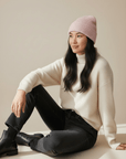 Eine Frau trägt eine LEY!-Wollmütze „Saha“ in Rosa und einen cremefarbenen Pullover; sie sitzt vor einem neutralen Hintergrund und lächelt.