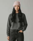 Eine Frau in LEY! Eine Wollmütze „Saha“ in Apricot, ein graues Sweatshirt und schwarze Jeans vor einem hellgrauen Hintergrund.
