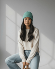Eine Frau mit einer LEY!-Wollmütze „Saha“ in Pastellgrün und einem weißen Pullover sitzt auf einem Hocker in einem sonnendurchfluteten Raum mit grauen Wänden.