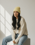 Eine Frau in einem LEY! „Saha“-Unisex-Wollmütze in Lila, einem cremefarbenen Pullover und Jeans sitzt auf einem Würfel in einem strahlend weißen Raum.
