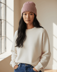 Eine Frau in einem cremefarbenen Pullover und einer LEY!-Wollmütze „Saha“ in Pink steht mit den Händen in den Taschen vor einem sonnendurchfluteten Fenster.