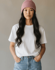 Eine Frau trägt eine LEY!-Wollmütze „Saha“ in Rosa, ein weißes T-Shirt und Jeans, die Hände in den Taschen, vor einem neutralen Hintergrund.