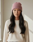 Eine Frau in LEY! Sie trägt die Wollmütze „Saha“ in Rosa, einen cremefarbenen Pullover und Jeans und steht in einem Innenraum, wobei natürliches Licht auf ihr Gesicht fällt.