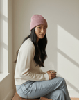 Eine Frau trägt die LEY!-Wollmütze „Saha“ in Rosa und einen weißen Pullover und sitzt mit einer Tasse in der Hand auf einer sonnigen Bank.