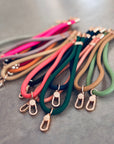LEY! Schlüsselanhänger aus farbenfrohen Stoffen mit goldenen Metallclips werden auf einer grauen Fläche in hellen und neutralen Farbtönen wie rosa, grün, orange, braun und schwarz präsentiert. Ideal als Lanyard Handy oder Handyband.