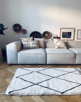 Ein modernes Wohnzimmer mit einem hellgrauen Sofa, gemusterten Kissen, runden Körben, gerahmter Kunst und dem BERBER TEPPICH - MAHIMA (1,50 x 2,50 m) von THE DANISH GIRL INTERIOR, einem weißen Wollteppich mit schwarzen geometrischen Linien auf Holzboden.