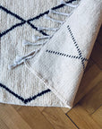 Eine Nahaufnahme des BERBER TEPPICH - MAHIMA (1,50 x 2,50m) von THE DANISH GIRL INTERIOR-geknüpft aus naturbelassener Wolle mit schwarzen Linien, Ecke gefaltet und Fransen sichtbar, auf Parkettboden.