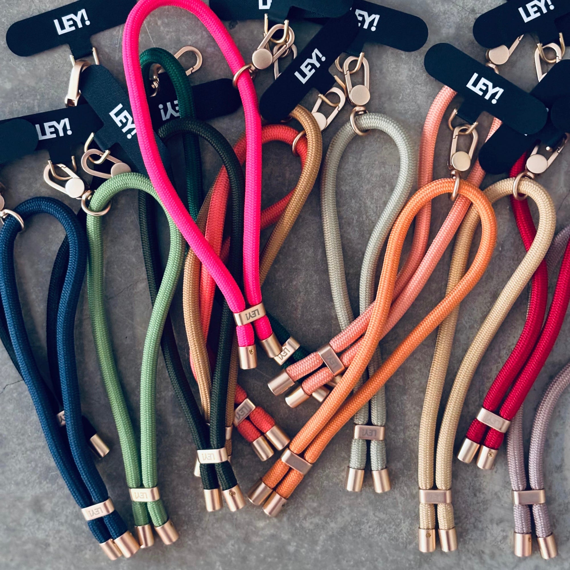 Eine Auswahl an bunten, individuell gestaltbaren LEY! Handykette / Schlüsselanhänger mit Metallclips ist auf einer grauen Oberfläche abgebildet, die jeweils ein schwarzes Etikett mit weißem LEY!-Branding aufweisen - ideal für Schlüssel oder als stylische Handyschlaufe.