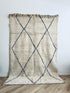 Der BERBER TEPPICH "MAHIMA" von LEY! (2,00 x 3,00m) ist ein cremefarbener, handgeknüpfter Teppich aus naturbelassener Wolle mit zwei großen schwarzen Rautenmustern und Oberkantenfransen, der aufrecht auf einem hellen Holzboden vor einer schlichten weißen Wand steht.